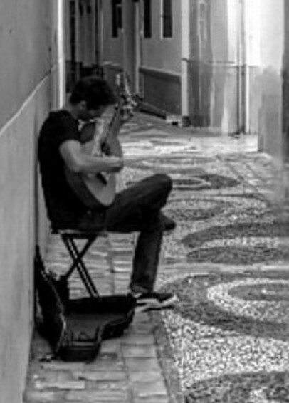 Zoom de la segunda foto. Guitarrista sentado, en primer plano. Viste de negro. Apoya la pierna derecha sobre la izquierda. Toca la guitarra con la cabeza gacha mirando a la propia guitarra.