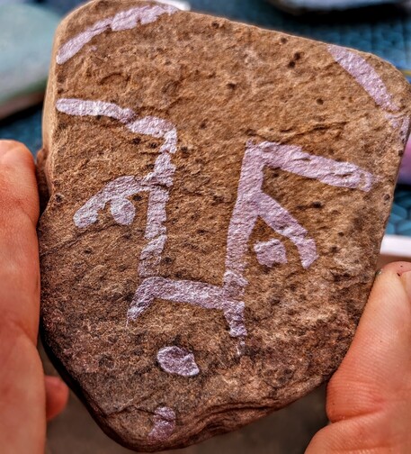 Piedra de color ocre con forma de cara, sobre la que se han pintado los rasgos de una cara de manera muy esquemática u con trazos más bien gruesos. La cara parece tener mirada triste.