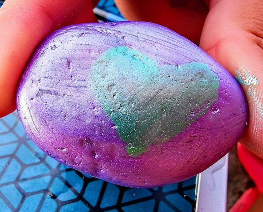 Típica piedra de la playa ovalada y de bordes suavizados y redondeados.
Pintada de color azulado y rosado metalizado brillante. En el centro hay pintado un corazón verde esmeralda metalizado.