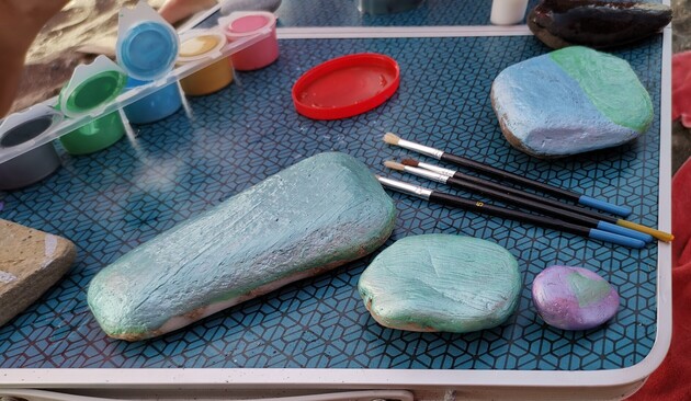 Mesita de playa donde se han realizado los trabajos y donde podemos ver piedras de playa, pinturas en pequeños botes tapados y unidos en ristra, y pinceles.