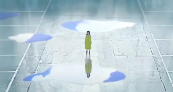 La protagonista de la película vestida con un chubasquero amarillo, en el centro de la imagen, en mitad de un aparcamiento rodeada de charcos de agua tras la lluvia.