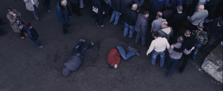 Dos hombres tirados en el suelo inmóviles son observados por una multitud de personas a su alrededor indiferente.