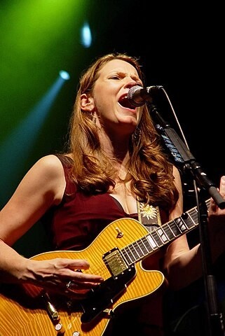 Foto en el escenario de una artista blanca y pelirroja tocando la guitarra y cantando con fuerza.
