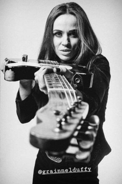 Foto de estudio en blanco y negro de la artista apuntando a la cámara con el mástil de su guitarra.
