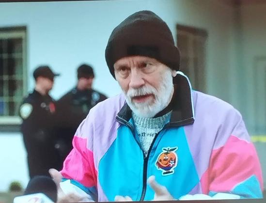 Un hombre mayor con barba blanca y un gorro negro está en primer plano, hablando. Lleva una chaqueta de chándal de colores brillantes en tonos pastel de rosa, turquesa y lavanda.  En la chaqueta tiene un parche con la imagen de naranjito.  De fondo, se ven dos policías ligeramente desenfocados.  La escena parece tener lugar en exteriores, frente a un edificio.