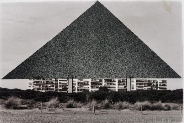 Un triangulo negro pintado con spray, sobre un bloque de apartamentos construidos en la playa. Se ve la arena en primer plano y algunas plantas.