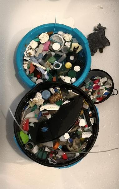 En un cubo de fregona d color negro se remojan trozos d plástico, tapas, cuerdas, etc. recogidos d la playa. Un barreño azul con más d estos elementos en remojo y un colador metálico negro con más plásticos. 