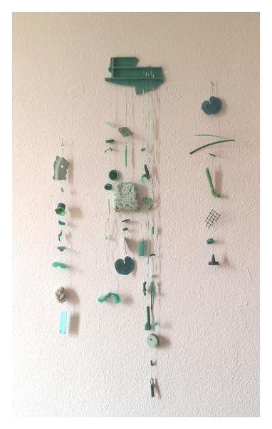 Foto frontal d un movil principal d siete hilos y dos móviles simples d un hilo a sus lados.Hechos con plásticos recogidos d la playa d tonos verdes.  De los hilos cuelgan palitos d chupete, tapas, un mechero, hojas plásticas, un auricular, etc.