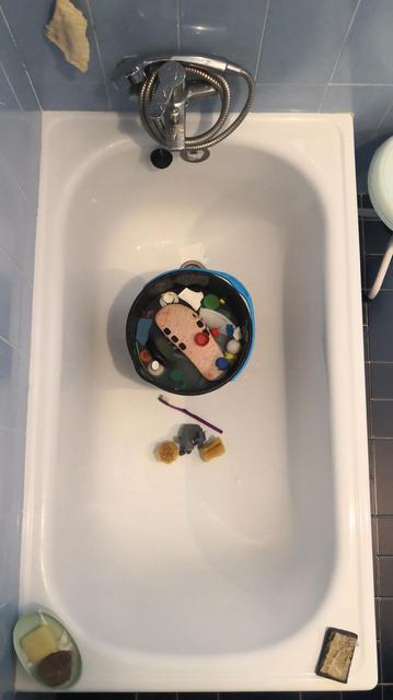 En una bañera blanca un cubo d fregona con plásticos varios recojidos d la playa están en remojo. Al lado del cubo un par d cepillos y esponjas para limpiarlos. 