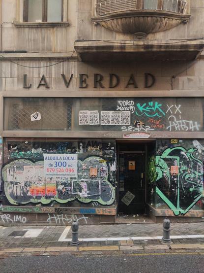 Fachada de el edificio La Verdad en la calle Capuchinas. El bajo comercial está cubierto de pintadas y de carteles publicitarios. Uno de ellos indica que se alquila. Sobre el escaparate hay un letrero que indica ”La verdad". Un poco más arriba se ve el inicio de un balcón decorado.