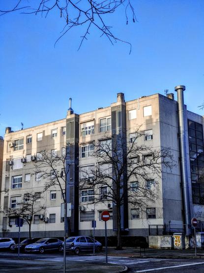 Una imagen de un edificio de apartamentos de varios pisos de color beige claro bajo un cielo azul despejado.  El edificio tiene muchos balcones y ventanas, y algunos árboles sin hojas se encuentran frente a él. Hay coches aparcados en la calle delante del edificio. Hay una señal de stop visible.  El edificio parece ser bastante moderno y está en una zona residencial.