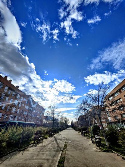 Un paseo arbolado se extiende hacia un cielo azul vibrante salpicado de nubes blancas y esponjosas.  A ambos lados del camino, hay bancos de parque y edificios de apartamentos de varios pisos de color marrón rojizo. Los árboles son mayoritariamente de hoja caduca y muestran sus ramas desnudas. Un autobús verde está estacionado cerca de uno de los edificios. El sol brilla intensamente, proyectando sombras largas en el pavimento. La escena transmite una sensación de calma y serenidad en un día soleado.