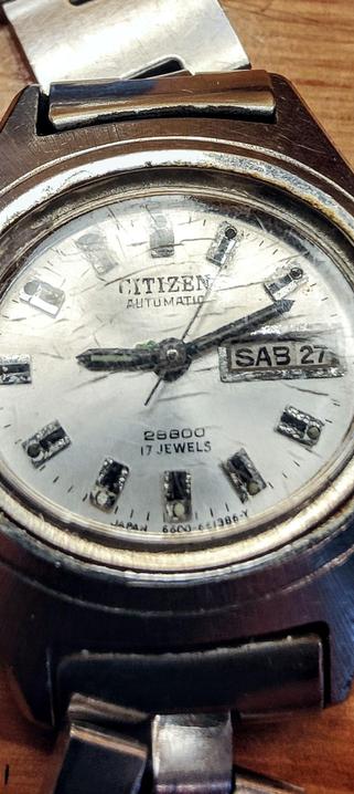 Una imagen de primer plano de un reloj de pulsera Citizen automático. La esfera es blanca con marcas negras y tiene una ventana de fecha que muestra "SAB 27".  El texto "CITIZEN AUTOMATIC" y "28800 17 JEWELS" también son visibles en la esfera. El reloj tiene una correa de metal oscura y muestra signos de uso y desgaste. El reloj descansa sobre una superficie de madera.