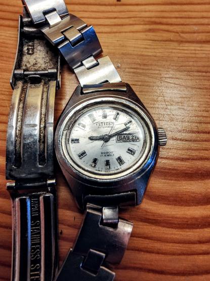 Un reloj Citizen automático de acero inoxidable, con una correa de eslabones también de acero inoxidable, descansa sobre una superficie de madera. El reloj muestra la fecha (Sábado 27) y tiene una esfera blanca con marcadores de hora de color negro. La correa del reloj parece estar un poco gastada o sucia. Una parte de la correa está separada del reloj, y se ve una inscripción que dice "Japan Stainless Steel".
