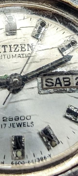 La imagen es un primer plano de un reloj Citizen automático. La esfera es de color blanco roto y muestra un ligero desgaste. Se puede leer claramente "CITIZEN AUTOMATIC", así como la fecha "SAB 2". También se ven las palabras "17 JEWELS" y un número que parece ser "28800". El cristal del reloj presenta algunas grietas. El diseño general del reloj es clásico y sencillo.
