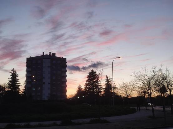 Una imagen de un edificio de apartamentos de varios pisos, silueteado contra un cielo crepuscular. El cielo muestra una mezcla de colores rosados y púrpuras, con nubes tenues dispersas.  Árboles de hoja caduca sin hojas, y algunos pinos, se encuentran en el primer plano, a los lados del edificio.  Un par de farolas iluminan la escena. El ambiente es tranquilo y sereno.