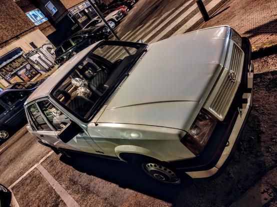 Una vista nocturna de un coche Opel Corsa blanco estacionado en un aparcamiento. El coche parece ser un modelo antiguo, con detalles como las molduras laterales y los faros. El interior del coche es visible a través de las ventanas, mostrando el salpicadero y algunos objetos en los asientos traseros. El coche está aparcado cerca de un edificio con grafitis en la pared, y otros coches estacionados se pueden ver en segundo plano, borrosos por la poca luz. La escena está iluminada por las luces de la calle y parece ser de noche.
