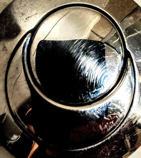 La imagen muestra un primer plano de un botón o perilla de metal circular, con un diseño de doble círculo concéntrico. La superficie metálica es oscura y brillante, con reflejos que sugieren un acabado pulido. El círculo interior es más oscuro y parece tener una textura ligeramente rayada o pulida, mientras que el círculo exterior es más claro y refleja algo de luz ambiental. La textura y el brillo sugieren que el objeto es metálico, posiblemente acero inoxidable o un metal similar. El conjunto crea una imagen abstracta, con contraste entre las zonas claras y oscuras del metal.