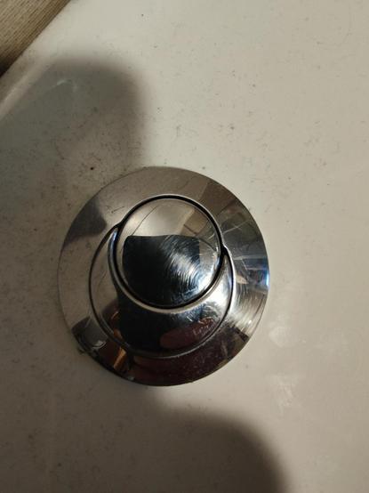 Imagen de un pulsador de cisterna de inodoro de metal cromado, redondo y con dos botones semicirculares. El pulsador está montado en una superficie blanca ligeramente sucia. Se ven algunos residuos o pelusas en la superficie blanca alrededor del pulsador.