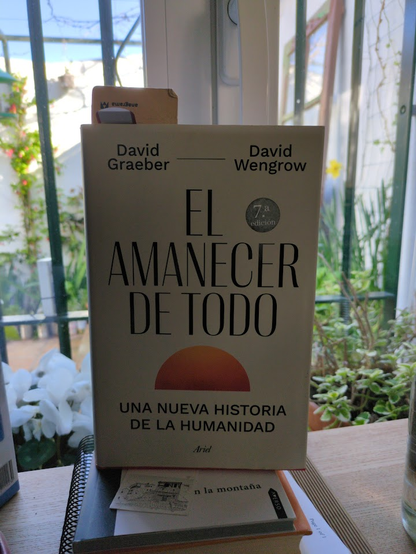 "El amanecer de todo" de Graeber & Wengrow en la ventana de mi escritorio. Afuera se ve un narciso, difuminado. 