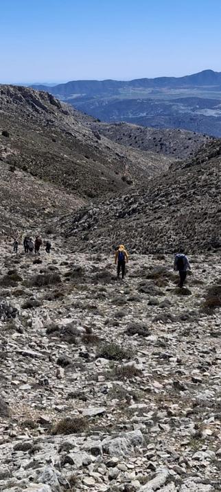 A group of hikers are trekking across a rocky, mountainous terrain (Sierra Maria, in Almería, Spain).  The hikers are spaced out along a path, with two individuals more prominent in the middle ground. The background shows a vast, distant landscape under a clear blue sky.