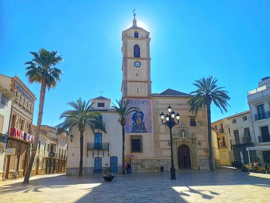 A sunny town square, dominated by a tall church tower with a clock.  Palm trees flank the church, which has a large, ornate entrance.  Low, white buildings line the square, and a street lamp stands in the center. A large poster is visible on the side of the church. This town is called Albox and is located in Almería, southern Spain.