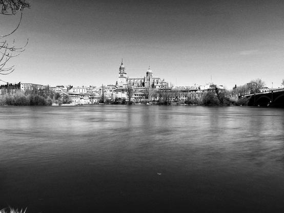Una foto en blanco y negro muestra una vista panorámica de un río tranquilo con una ciudad en el fondo.  En el centro de la imagen, se alza una imponente catedral o conjunto de edificios religiosos, con torres y cúpulas visibles.  A los lados, se extiende una línea de edificios de menor altura, característica de una ciudad histórica.  El río ocupa la mayor parte del primer plano, mostrando una superficie ligeramente ondulada. Un puente arqueado es visible en la parte derecha del fondo. La escena está bañada por una luz difusa, creando una atmósfera serena y pacífica.