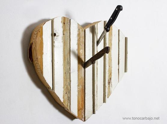 Un corazón construido con vigas recuperadas de pisos de desahucios bancarios, con dos cuchillos de cocina clavados l con mangos uno negro y el otro marrón.   El corazón está colgado en una pared blanca.