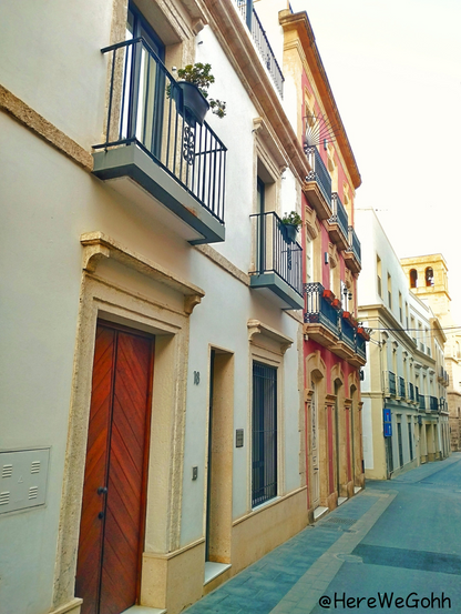 Una estrecha callejuela muestra una hilera de edificios de dos o tres pisos, de arquitectura española. Los edificios son de color blanco cremoso y un edificio destaca por sus tonos rojizos.  Las fachadas presentan balcones con barandillas de hierro forjado, algunos con macetas de plantas. Una puerta de madera oscura y veteada destaca en la planta baja de uno de los edificios. La calle está pavimentada. Es un día despejado y soleado, como.es habitual en Almería.